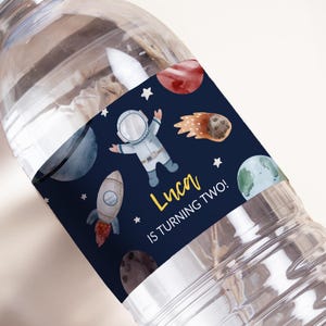 Puede incluir: Una botella de agua con una etiqueta personalizada con un astronauta, planetas, un cohete y el texto "Luca IS TURNING TWO!". El diseño está sobre un fondo azul oscuro con estrellas blancas.