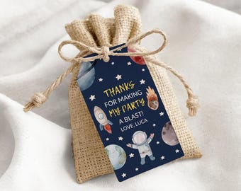 May include: A navy blue gift tag with a space theme, featuring a rocket, planets, stars, and an astronaut. The tag reads "Thanks for making my party a blast! Love, Luca" and is attached to a burlap gift bag with a drawstring closure.