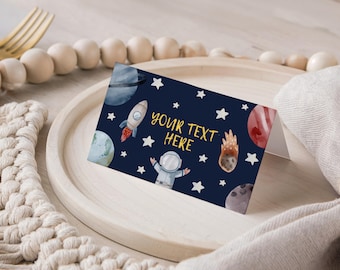 以下が含まれることがあります： 水彩画の宇宙をテーマにした折り畳みカード。濃い青色のカードには、ロケット、惑星、星、宇宙飛行士が描かれています。「YOUR TEXT HERE」の文字は黄色です。カードは木製のトレイに置かれています。