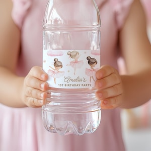 May include: Clear plastic water bottle with a label featuring three ballerina illustrations in pink tutus. The label also includes the text "Amelia's 1st Birthday Party". The bottle is held by a person wearing a pink dress.