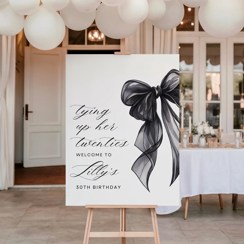 May include: A white sign on a wooden easel with a black bow illustration and text that reads "Tying up her twenties, Welcome to Lilly's, 30th Birthday." White balloons are suspended above the sign.