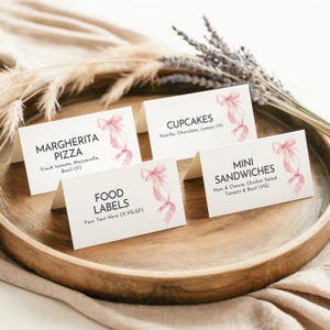 May include: Four white folded cardstock food labels featuring a watercolor pink ribbon illustration. The labels display text including MARGHERITA PIZZA, CUPCAKES, MINI SANDWICHES, and FOOD LABELS with various ingredient descriptions and dietary markers.