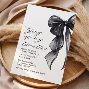 Pu&ograve; includere: Un biglietto d'invito bianco con il testo "Tying up my twenties" e i dettagli dell'evento. Una grande ed elegante illustrazione di fiocco nero adorna il lato destro. Il biglietto &egrave; appoggiato su un vassoio di legno con piante decorative essiccate.