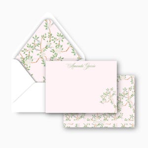 May include: A stationery set featuring a light pink card and envelope with a floral design. The envelope interior and the back of the card have a repeating pattern of white flowers and green leaves on brown branches. The card is printed with the name "Amanda Garcia" in green.