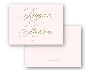 May include: Two light pink stationery cards with the names "Reagan Morton" in elegant green script. The bottom card has the text "@riverpaperco" in a smaller font.