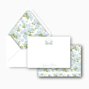 May include: White stationery set featuring a floral hydrangea design. The set includes an envelope with a floral interior, a blank card with a floral crest, and a matching patterned card. The name "Reagan Morton" is printed on the card.