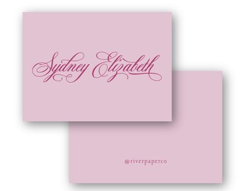 May include: Two light pink stationery cards. The top card features the name "Sydney Elizabeth" in a cursive, burgundy font. The bottom card has the text "@riverpaperco" in a smaller, burgundy font.