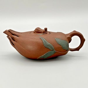 May include: A reddish-brown teapot with a unique design. The teapot is shaped like a hand with leaves and a small knob on the lid. The teapot is made of clay and has a rustic look.