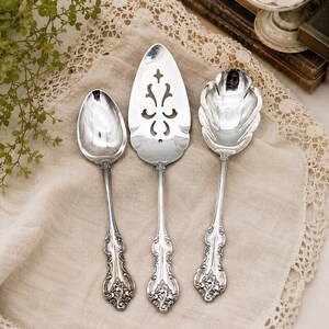 May include: A set of three silver-toned serving utensils featuring ornate, embossed floral handles. The collection includes a solid oval spoon, a pointed cake server with a central decorative cutout, and a scalloped-edge serving spoon.