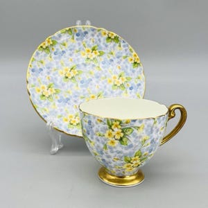 Pode incluir: Conjunto de chá floral. A chávena e o pires apresentam um fundo azul claro com padrões florais amarelos e brancos. A chávena tem uma pega e base douradas, e o pires tem uma borda ondulada.