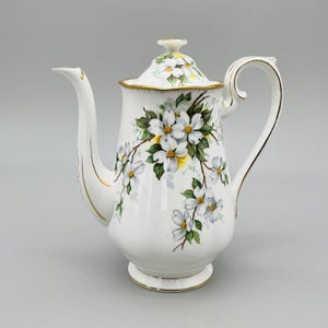 Puede incluir: Cafetera de cerámica blanca con tapa, con un diseño floral de flores blancas con centros amarillos y hojas verdes. La cafetera tiene un borde y un asa dorados, y un pico largo.