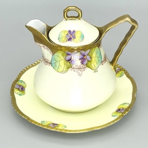 May include: Cream-colored porcelain creamer and saucer set. The creamer has a gold handle and trim, with a lid. Both pieces are decorated with purple violets and green leaves. The saucer has a scalloped edge and gold trim. A charming floral design.