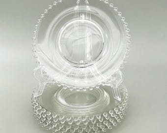 May include: Clear glass plates with a beaded edge. The plates are stacked, with one plate displayed on a clear plastic plate stand. The plates have a simple, elegant design, suitable for serving appetizers or desserts.