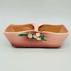 Puede incluir: Una jardinera de cerámica rosa con un interior dividido y un asa. El exterior presenta un diseño floral con hojas verdes y flores blancas y amarillas. La jardinera tiene forma rectangular.