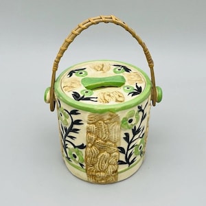 May include: A ceramic jar with a green and brown floral design and a bamboo handle. The jar has a lid with a small hole in the center.