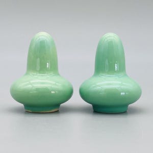 May include: Two matching, light green ceramic bottle stoppers. Each stopper has a rounded base and a long, tapered top. The glossy finish reflects light, highlighting the smooth curves and subtle color variations.