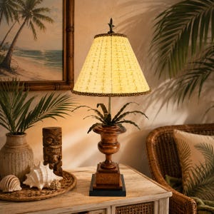 May include: A table lamp with a dark brown wooden base shaped like a pedestal, topped with a conical cream-colored lampshade featuring a repeating star pattern and beaded trim. A metal finial sits at the peak of the shade, and dark green foliage.
