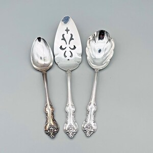 May include: Three pieces of ornate silver serving cutlery are displayed against a gray background. The set includes a spoon, a pie server with a decorative cutout, and a serving spoon. Each piece has a detailed floral design on the handle.
