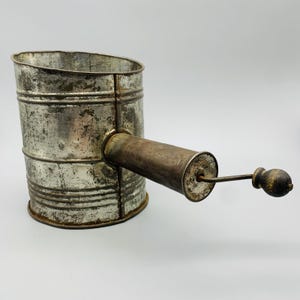 Op de afbeelding: Vintage metalen bloemzeef met een cilindrisch lichaam en een houten handvat. De zeef heeft een verweerde, verouderde uitstraling, met zichtbare slijtage op het metalen oppervlak. De handgreep is cilindrisch en aan de zijkant bevestigd.