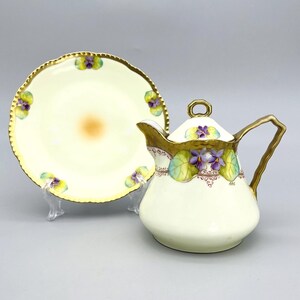May include: A cream-colored porcelain creamer and saucer set. The set features hand-painted purple violets and green leaves with gold trim. The saucer is round with a scalloped edge. The creamer has a curved handle and a lid with a gold ring.