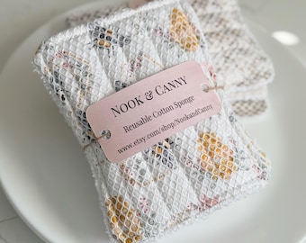 May include: A white cotton sponge with a floral pattern in yellow and pink. The sponge is labeled "NOOK & CANNY Reusable Cotton Sponge" and includes the website address "www.etsy.com/shop/NookandCanny".
