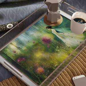 Puede incluir: Una bandeja de servicio de acrílico transparente con un estampado floral colorido, que contiene una taza de café, una cafetera plateada y un mando a distancia. Un smartphone y un libro abierto están cerca. La bandeja tiene asas plateadas.