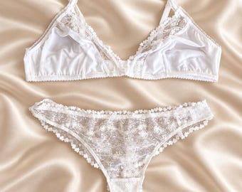 May include: A white lingerie set consisting of a soft-cup bralette and a matching thong. The bralette features delicate lace trim along the neckline and adjustable shoulder straps. The thong is crafted from intricate floral lace with scalloped edges.