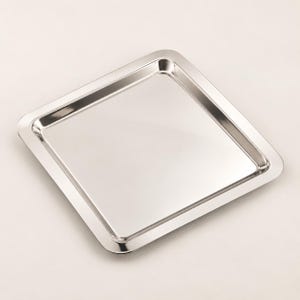 Pode incluir: Uma bandeja quadrada de metal prateado polido com uma superfície lisa e reflexiva e cantos arredondados. A bandeja apresenta uma borda ligeiramente elevada que emoldura o centro plano, criando um visual limpo com acabamento metálico.
