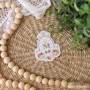Puede incluir: Una pegatina blanca con un dise&ntilde;o floral y el texto "Oh Mar&iacute;a, concebida sin pecado, ruega por nosotros que recurrimos a ti." La pegatina presenta una cruz y la letra "M" en el centro.