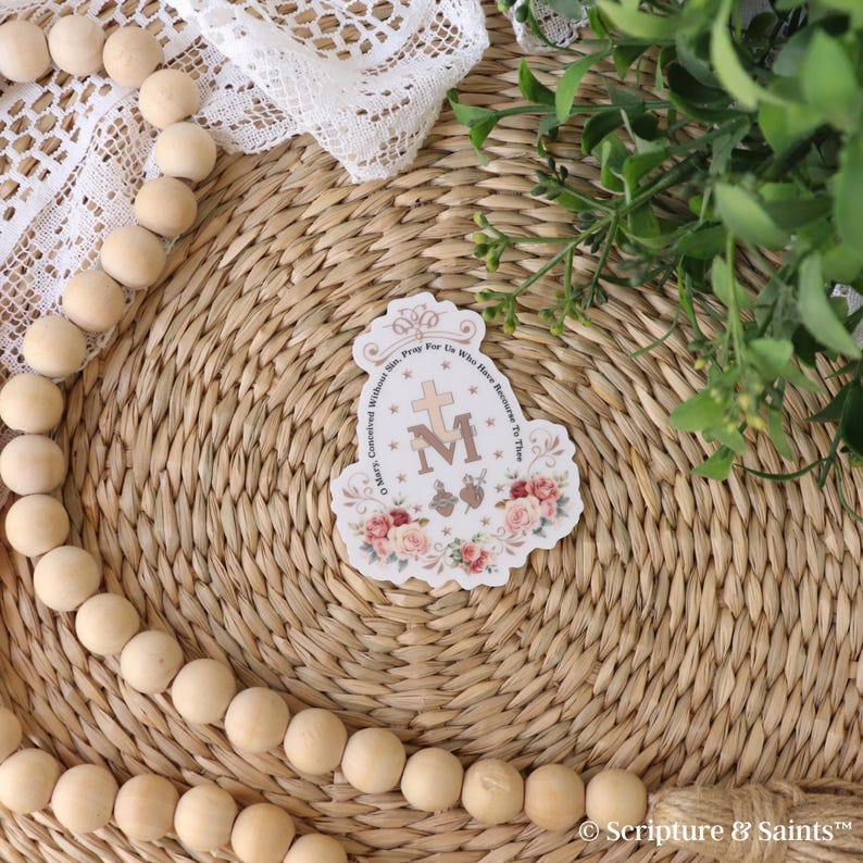 Puede incluir: Una pegatina blanca con un dise&ntilde;o floral y el texto "Oh Mar&iacute;a, concebida sin pecado, ruega por nosotros que recurrimos a ti." La pegatina presenta una cruz y la letra "M" en el centro.