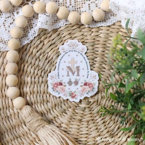 Puede incluir: Una pegatina blanca con un dise&ntilde;o floral y el texto "Oh Mar&iacute;a, concebida sin pecado, ruega por nosotros que recurrimos a ti" y una "M" estilizada con una cruz y un coraz&oacute;n.