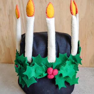 May include: A black fabric Christmas decoration shaped like a cake, adorned with white felt candles with yellow and orange flames. A green felt holly garland with red berries encircles the base, creating a festive holiday theme.