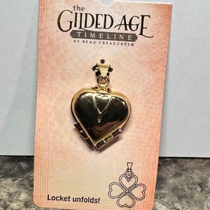 May include: A gold-colored heart-shaped locket on a pink card. The card reads "the Gilded Age Timeline" and "Locket unfolds!" The locket has a small loop at the top for a chain. A clover design is also on the card.