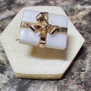May include: A white rectangular ring with a gold bow and gold band detail. The ring is set on a white and beige surface, with a dark, textured background. The bow is intricately designed, adding a touch of elegance to the jewelry.