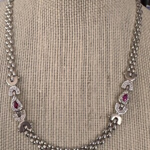 May include: A silver chain necklace with two teardrop-shaped pendants. Each pendant is set with a small, red gemstone and surrounded by smaller clear gemstones.