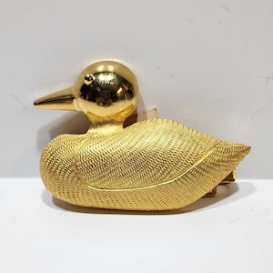 May include: A gold-colored duck brooch with a textured body and smooth head. The duck has a pointed beak and a small eye detail. The brooch is a vintage accessory.
