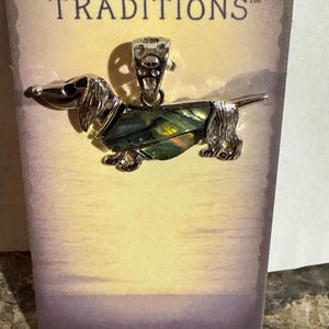 May include: A silver-toned dachshund pendant with an iridescent abalone shell body. The pendant is on a card with the word "TRADITIONS" and a scenic background.