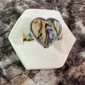 May include: A silver ring with a heart-shaped abalone shell inlay. The iridescent shell features shades of blue, green, and gold. A small green gemstone is set on the band. The ring is displayed on a white hexagonal stand.