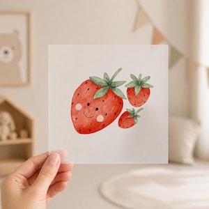 Könnte beinhalten: Aquarellillustration von drei Erdbeeren mit grünen Blättern. Die größte Erdbeere hat ein lächelndes Gesicht mit rosigen Wangen. Die Erdbeeren sind rot mit kleinen schwarzen Samen, und das Bild wird von einer Hand gehalten.