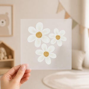 Könnte beinhalten: Ein quadratisches, durchscheinendes Blatt mit drei weißen Gänseblümchen-Illustrationen mit gelben Zentren. Die Gänseblümchen sind in einer Gruppe angeordnet, wobei das Blatt von einer Hand gehalten wird. Der Hintergrund ist eine weiche, neutrale Farbe.