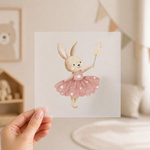 Könnte beinhalten: Aquarell-Illustration eines Hasen in einem rosa Kleid mit weißen Blumenakzenten, der einen sternförmigen Zauberstab hält. Der Hase hat hellbraunes Fell und lange Ohren. Das Bild befindet sich auf einer transparenten, quadratischen Oberfläche.
