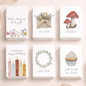 Könnte beinhalten: Ein Stapel von sechs weißen Grußkarten mit Aquarellillustrationen. Designs umfassen Blumen, einen Umschlag, Pilze, Kerzen, einen Kranz und einen Cupcake. Jede Karte hat eine andere Nachricht auf Deutsch.