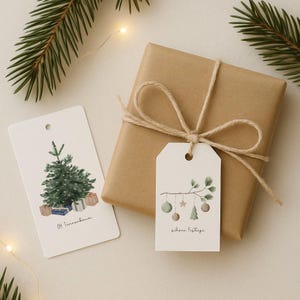 Könnte beinhalten: Ein Geschenk in braunes Papier verpackt und mit Bindfaden verschnürt, begleitet von zwei Geschenkanhängern. Ein Anhänger zeigt einen Aquarell-Weihnachtsbaum und den Text "Oh Tannenbaum". Der andere Anhänger zeigt einen Zweig mit Ornamenten und dem Text "Schöne Festtage."