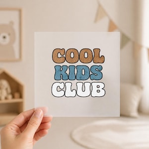 Könnte beinhalten: Ein quadratischer, transparenter Druck mit dem Text "COOL KIDS CLUB" in einer Retro-Schriftart. Das Wort "COOL" ist braun, "KIDS" ist blau und "CLUB" ist weiß, alle mit schwarzen Umrissen. Der Druck wird von einer Hand gehalten.