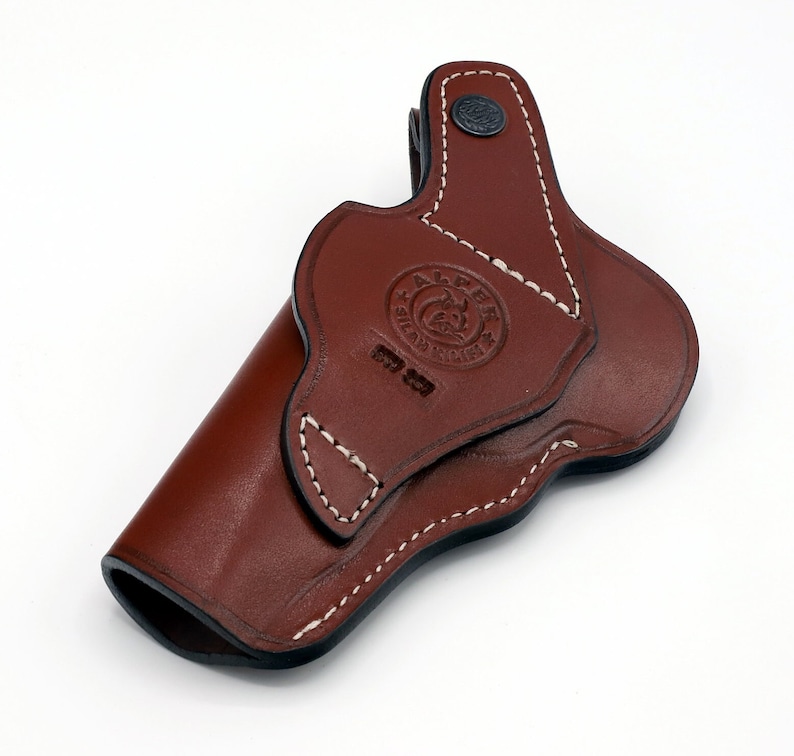 May include: A brown leather holster with white stitching and a black button. The holster has a logo that says "ALPS" and "SILAS BROTHERS".