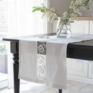 May include: White linen table runner with a delicate lace trim. The runner is on a black table with a vase of white flowers.
