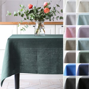 May include: A green linen tablecloth with a subtle textured pattern, displayed on a table in a kitchen setting. A vase with pink roses and greenery sits on the table. The image also shows a variety of other tablecloth colors.