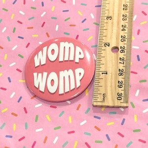 Può includere: Un bottone rosa con il testo "WOMP WOMP" in lettere bianche. Il bottone ha un diametro di circa 5 cm.