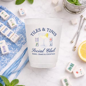 May include: A frosted plastic cup with the text "TILES & TINIS Social Club" and a cocktail illustration. The image includes Mahjong tiles, mint sprigs, lemon slices, sugar cubes, and a bucket of ice.