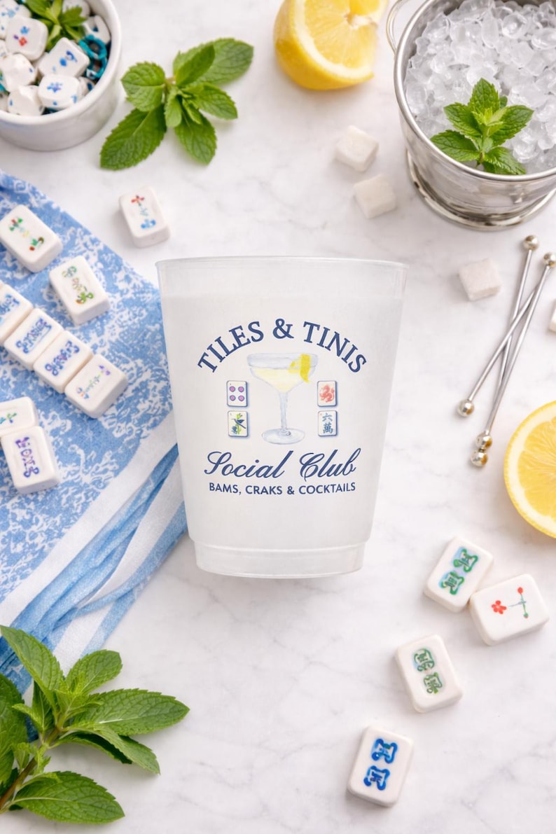 May include: A frosted plastic cup with the text "TILES & TINIS Social Club" and a cocktail illustration. The image includes Mahjong tiles, mint sprigs, lemon slices, sugar cubes, and a bucket of ice.